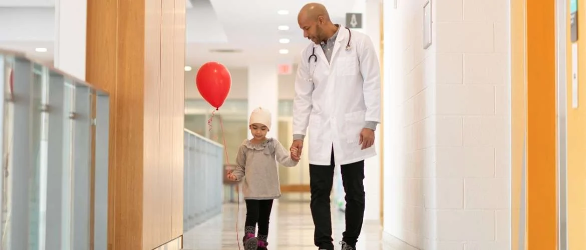 Em um corredor claro de hospital, uma menina pequena com gorro na cabeça segura um balão vermelho e anda de mãos dadas com um homem de jaleco branco e estetoscópio no pescoço.