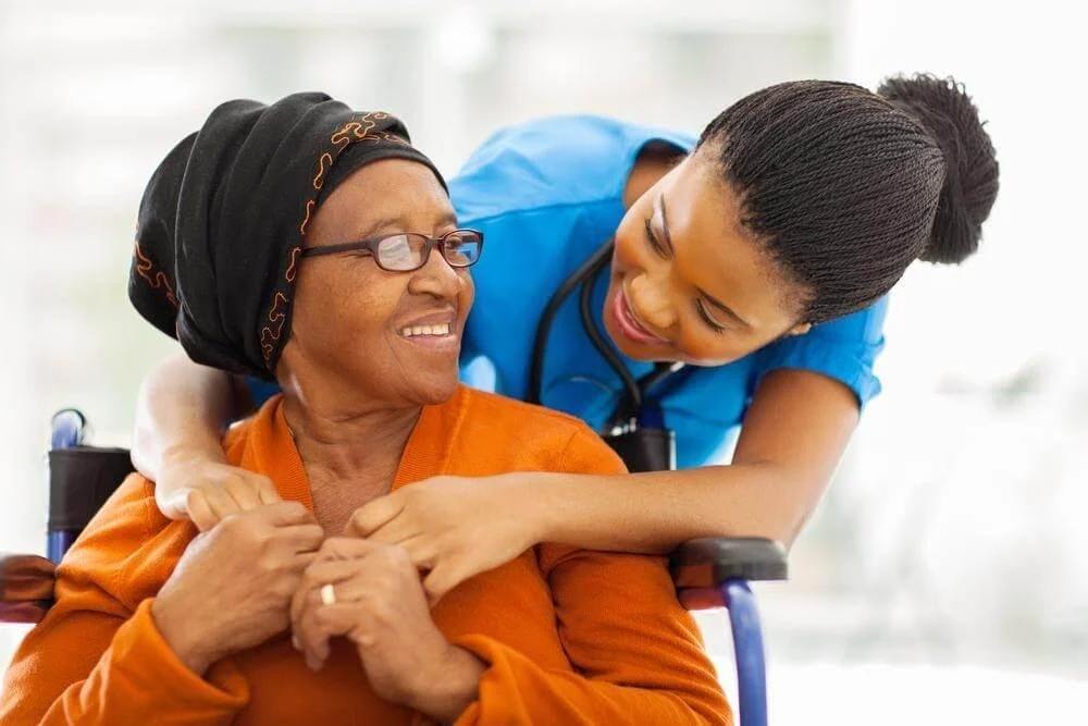 Uma mulher idosa negra está sentada em uma cadeira de rodas sorrindo, recebe um abraço de uma médica negra com jaleco azul e com um medidor de pressão no pescoço.