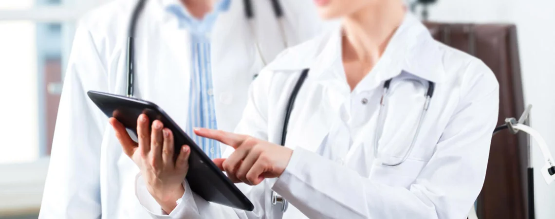 O tronco de mulher com jaleco branco aparece, ela tem o estetoscópio no pescoço, segura um tablet em que mostra algo para outro médico homem com jaleco branco e estetoscópio no pescoço.